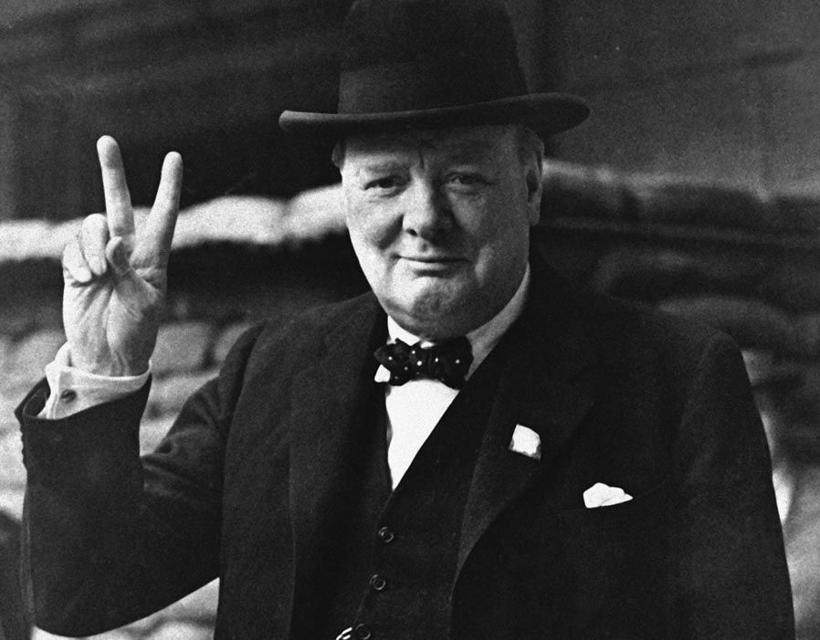 A man in a suit and hat making the peace sign.